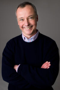 Chris Kenber in a dark blue jumper and a shirt, smiling with his arms crossed