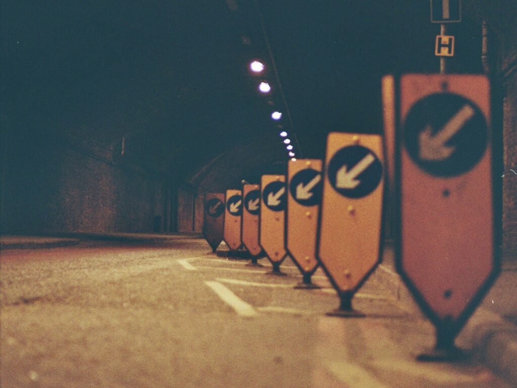 A row of street signs on the side of the road pointing down to the left, narrowing the lane